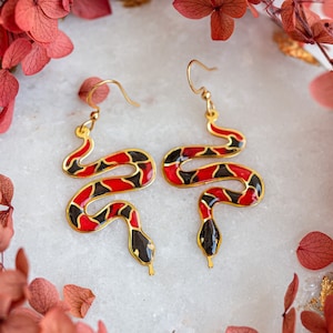 May include: A pair of gold snake earrings with red and black enamel. The snakes are curled up and have a detailed design.
