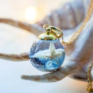 May include: A gold chain necklace with a glass orb pendant. The orb contains a white starfish, a seashell, and blue sand.