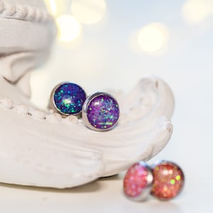 Peut inclure: Boucles d'oreilles à clous avec un design scintillant et pailleté. Les boucles d'oreilles ont une forme ronde et bombée avec une lunette de couleur argentée. Les couleurs principales sont des nuances de violet, de bleu et de vert. D'autres boucles d'oreilles roses et oranges sont visibles.
