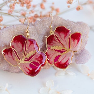 Może przedstawiać: Para złotych kolczyków z motywem czerwonych i różowych kwiatów. Kwiaty mają białe środki i złote akcenty.