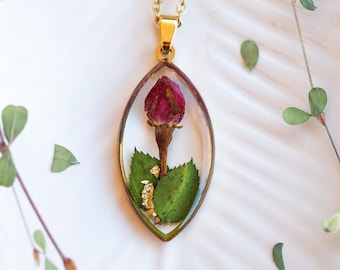 May include: A gold-colored necklace with a pendant containing a preserved red rose bud, green leaves, and gold flakes. The pendant is an oval shape with a gold-colored frame. The necklace chain is also gold-colored.