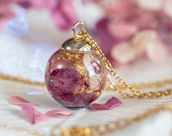 May include: A gold chain necklace with a round pendant featuring dried pink flowers and gold leaf encased in clear resin.