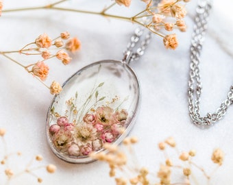 Puede incluir: Un collar de cadena de plata con un colgante ovalado que presenta flores rosadas y blancas prensadas encapsuladas en resina transparente.
