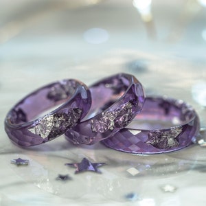 May include: Three faceted, purple resin rings with silver leaf accents. The rings are displayed on a reflective surface with star-shaped confetti. The rings are from the brand 13th Psyche.
