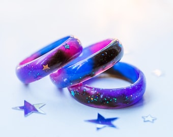 Puede incluir: Tres anillos de resina con temática de galaxia con purpurina y estrellas incrustadas. Los anillos están apilados uno encima del otro, con el anillo superior azul y morado, el anillo del medio rosa y morado, y el anillo inferior morado y azul.