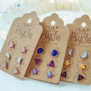 Può includere: Tre set di piccoli orecchini colorati e scintillanti su cartoncini marroni. Ogni cartoncino riporta il testo "13th Psyche" e un indirizzo web. Gli orecchini hanno la forma di stelle, lune e triangoli.