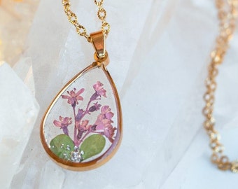 Può includere: Una collana con catena dorata con un ciondolo a forma di lacrima con un motivo di fiori pressati. Il ciondolo è pieno di fiori rosa e verdi ed è posto su uno sfondo di cristallo di quarzo bianco.