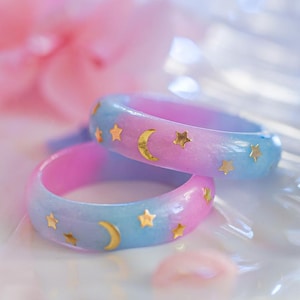 Könnte beinhalten: Zwei Harzringe mit einem Farbverlauf aus Rosa, Lila und Blau. Jeder Ring ist mit goldenen Halbmonden und Sternen verziert. Die Ringe werden auf einem weichen, verschwommenen Hintergrund mit rosa und blauen Blumen präsentiert.