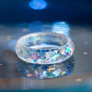 Könnte beinhalten: Ein klarer Harzring mit eingebettetem schimmerndem Glitzer. Der Ring liegt auf einer blauen Oberfläche.