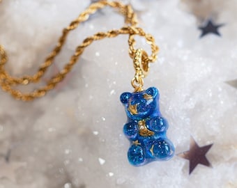 May include: A gold chain necklace with a blue gummy bear charm. The charm is sparkly and has a gold crescent moon on its chest.