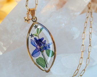 May include: A gold chain necklace with a clear resin pendant featuring a pressed purple flower and green leaves. The pendant is shaped like a teardrop and hangs from a delicate gold chain.