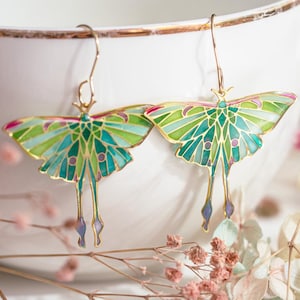 Puede incluir: Un par de pendientes dorados con un diseño de polilla Luna. Los pendientes tienen alas verdes y azules con detalles intrincados, acentuados con rosa y morado. Los pendientes se muestran frente a un cuenco blanco.