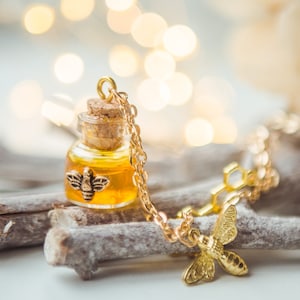 Può includere: Una collana d'oro con un piccolo ciondolo a forma di bottiglia di vetro riempita di miele e un ciondolo a forma di ape. La collana è su una superficie di legno con uno sfondo sfocato di luci.