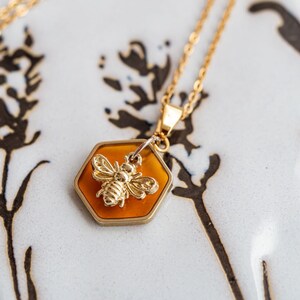 Può includere: Collana con catena dorata e ciondolo esagonale con un'ape dorata su sfondo ambrato.