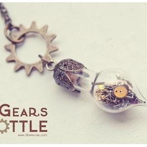 以下が含まれることがあります： 歯車の形をしたペンダントが付いた銀色のネックレス。ペンダントは、小さな歯車やその他の時計部品で満たされた透明なガラス瓶です。ペンダントの下には、「GEARS BOTTLE」と「www.13thpsyche.com」の文字が印刷されています。