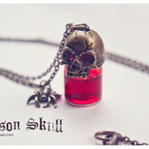 May include: A silver chain necklace with a glass vial pendant topped with a brass skull. The vial is filled with a red liquid. The text "Poison Skull" is printed below the pendant.