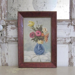 May include: An oil painting in a dark red wooden frame depicting a bouquet of yellow, pink, and orange flowers arranged in a blue vase. The flowers sit on a white cloth surface against a light gray background. The artist signature is visible.