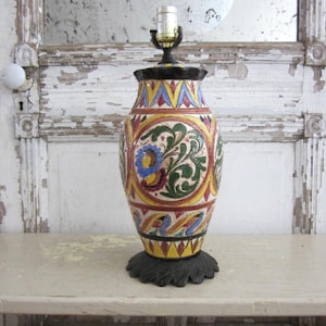 May include: A decorative ceramic table lamp with a colorful, ornate design. The lamp features a black base and top, with a central body adorned with floral and geometric patterns in yellow, blue, red, and green. The lamp is topped with a standard light socket.