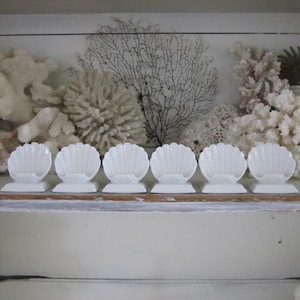 May include: Six white seashell-shaped dishes are arranged in a row. The dishes are on a shelf with coral and other decorative elements in the background. The overall theme is coastal decor.