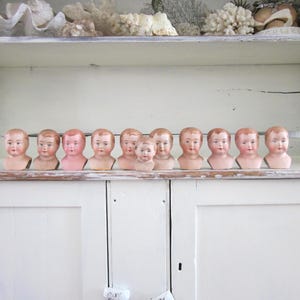 May include: A collection of antique doll heads with painted features, arranged on a shelf. The doll heads have a porcelain appearance with pink cheeks and rosy lips. The shelf is part of a white cabinet, with decorative items on the top shelf.