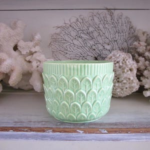 May include: A light green ceramic planter with a leaf pattern. The planter has a ribbed top and a series of repeating leaf shapes around the sides. The planter is sitting on a distressed wooden surface.