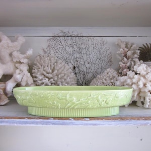 May include: A light green ceramic serving dish with a detailed floral pattern. The dish is oval-shaped and sits on a raised base. The background features white coral and other decorative elements, creating a coastal theme.