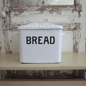 May include: A white bread box with a lid and handle, featuring the word "BREAD" in bold black letters. The box is rectangular and sits on a light-colored surface, with a distressed white door in the background.