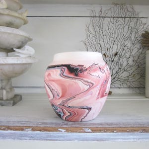 May include: A small, round, pink and black marbled ceramic pot with a white background. The pot is sitting on a white shelf.