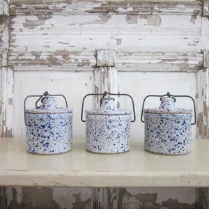 May include: Three cylindrical ceramic crocks with lids, each featuring a white base with a blue speckled pattern. Each crock includes a dark metal wire handle attached to the lid, allowing for a secure closure and easy transport of the containers.