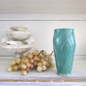 May include: A teal ceramic vase with a leaf design sits on a white shelf with a white alabaster fountain and a bunch of yellow grapes.