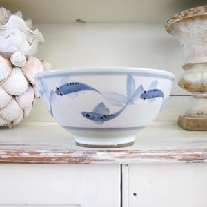 May include: White ceramic bowl with a blue fish design. The bowl has a rounded shape and a slightly flared rim. The fish are stylized with blue bodies and fins. The bowl is sitting on a white shelf.