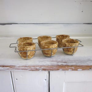 May include: Six small, round, ceramic bowls with a speckled tan and brown pattern, held in a silver wire rack. The bowls are arranged in two rows of three. The background is a white, distressed wooden surface.