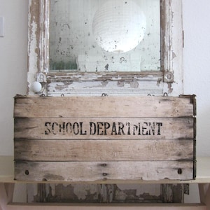 May include: A rustic wooden crate with the words "SCHOOL DEPARTMENT" stenciled in black. The crate is placed in front of a distressed, antique mirror with peeling white paint. A white paper lantern hangs in the reflection.