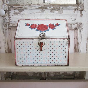 May include: A white metal bread box with a slanted lid featuring a red rose floral design. The front panel displays a repeating pattern of small red and blue diamond shapes and includes a central red latch handle for opening the container.