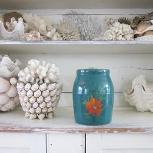 May include: A teal ceramic jar with a hand-painted orange flower design sits on a white shelf next to a white shell-covered planter filled with white coral. Other white decorative shells and coral pieces are arranged on the shelf above and below.