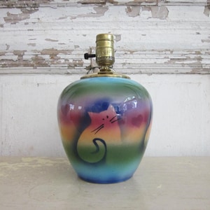 May include: A ceramic table lamp with a colorful, gradient design. The lamp features a cat illustration in pink, yellow, and green. The lamp has a brass-colored socket and a black on/off switch. The base is a rounded, bulbous shape.