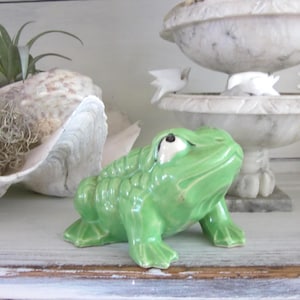 May include: A ceramic frog figurine in a glossy green finish. The frog has white eyes and is posed in a crouching position. The figurine is on a white surface, with a seashell and a decorative marble urn in the background.