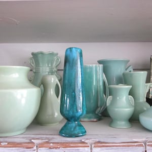May include: A collection of ceramic vases in various shades of blue and green. The vases vary in shape and size, including a tall, turquoise vase and several rounded and handled designs. The vases are arranged on a white shelf.