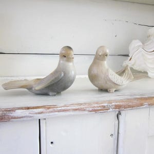 May include: Two ceramic bird figurines in shades of gray and beige sit on a white surface. The birds have detailed wings and are positioned facing different directions. A large, white, shell-like object is partially visible in the background.