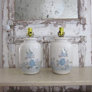 May include: Two matching white ceramic table lamps. Each lamp features a blue rose design and a gold-colored lamp holder. The lamps are displayed on a light-colored shelf against a distressed white background.