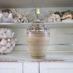 May include: A ceramic table lamp with a beige and brown textured pattern. The lamp has a brass-colored socket and a neutral color palette. The background features white shelves with seashell decor.