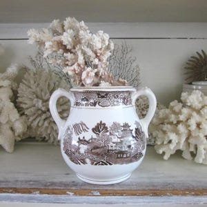 May include: A white ceramic sugar bowl with two handles and a brown transferware design depicting a landscape scene. The bowl is filled with coral and is displayed on a shelf with other coral pieces.