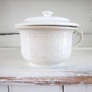 May include: A white ceramic chamber pot with a lid and handle. The pot has a scalloped design around the body and a small knob on the lid. The chamber pot is sitting on a white wooden surface.