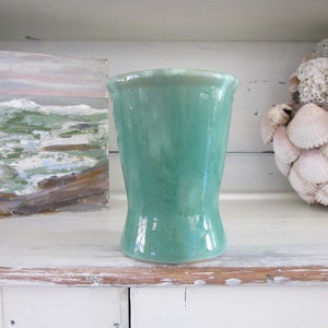 May include: A teal green ceramic vase with a flared rim and a slightly pinched waist. The vase is sitting on a white shelf with a painting and seashells in the background.