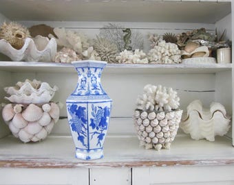 May include: A hexagonal ceramic vase featuring a white base with blue floral and botanical patterns. The vase has a flared rim and a tapered neck, standing prominently among various white seashells and coral specimens on a white shelf.
