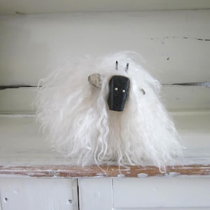 May include: A handmade sheep figurine with a fluffy white wool body and a black face. The sheep has small, upright ears and two small, black eyes. The figurine is sitting on a white shelf.