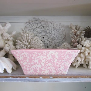 May include: A rectangular pink planter with a white, textured pattern. The planter is displayed on a shelf with white coral, seashells, and other decorative elements. The overall theme is coastal decor.
