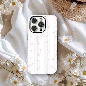 Può includere: Custodia per telefono bianca con motivo floreale e a nastro. La custodia presenta un delicato design di fiori rosa e nastri su sfondo bianco a righe verticali. La custodia del telefono ha un modulo fotocamera nero.