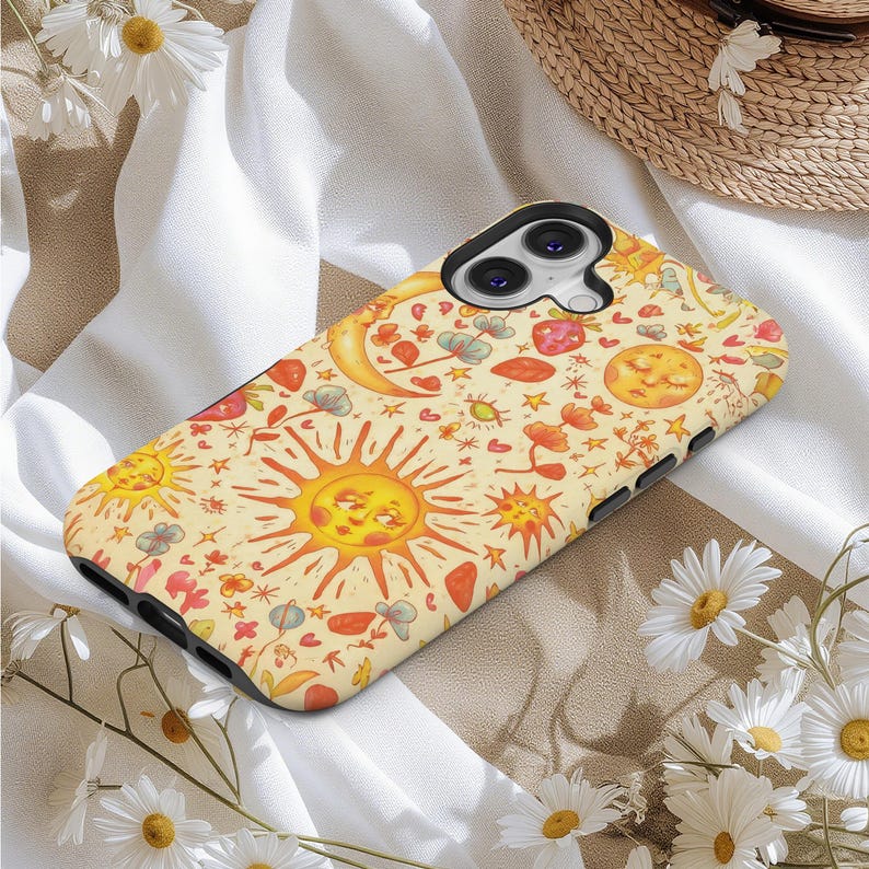 May include: A phone case with a whimsical design featuring smiling suns, moons, stars, and floral elements in shades of yellow, orange, and red. The case has a black border and is set against a backdrop of white fabric and daisies.