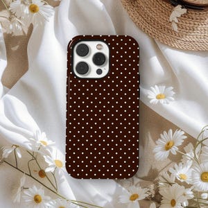 May include: A brown phone case with a white polka dot pattern. The case has a black border and a camera cutout. The phone case is surrounded by white daisies and a straw hat.